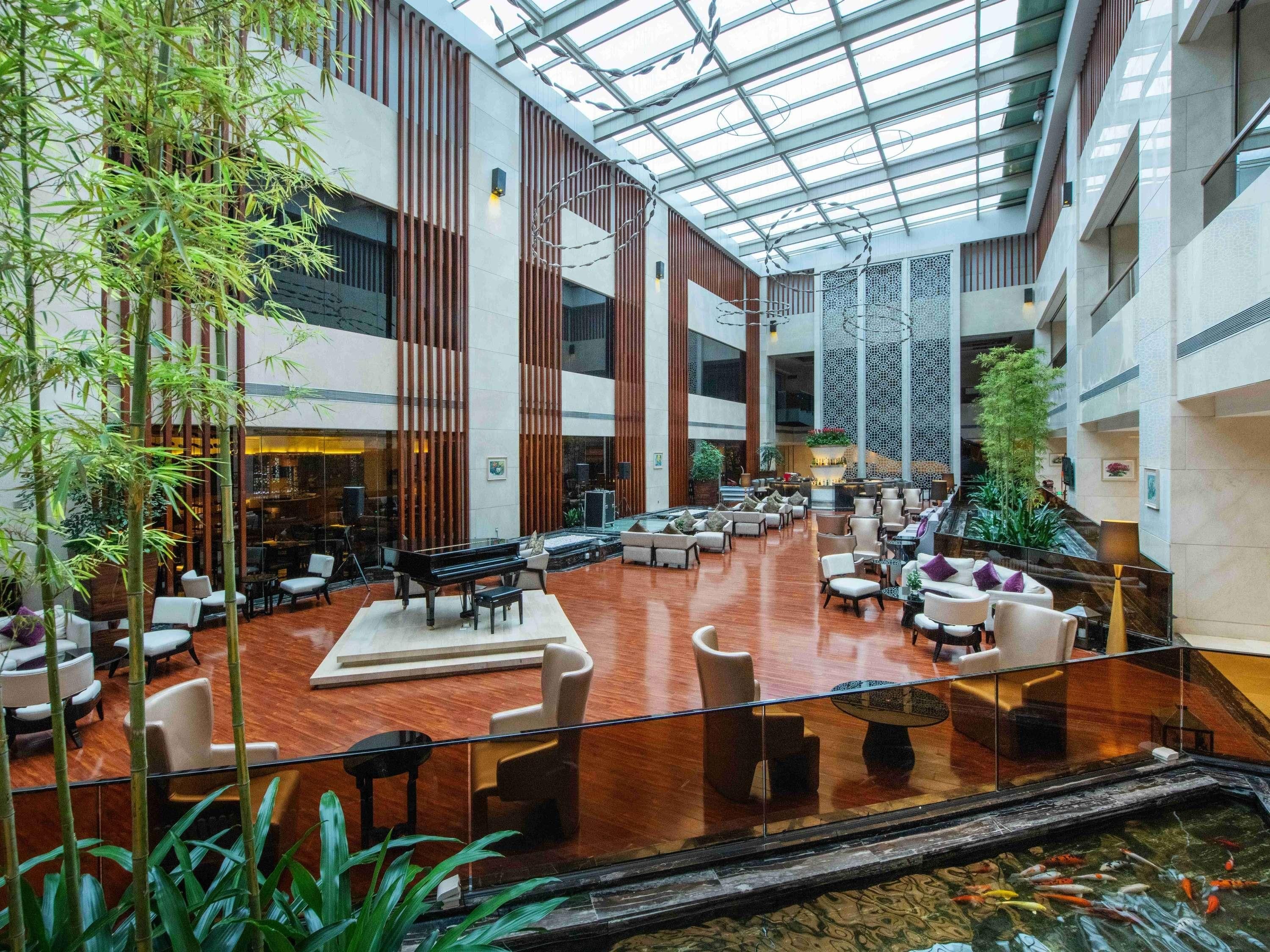 A spacious indoor atrium with a glass ceiling, seating areas, a piano, and a koi pond.