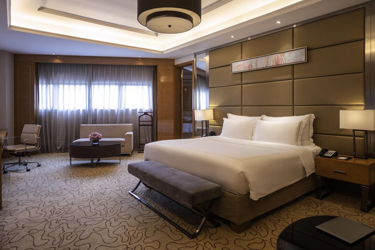 A hotel room with a large bed, a desk, a chair, a bench, a lamp, and a bedside table.