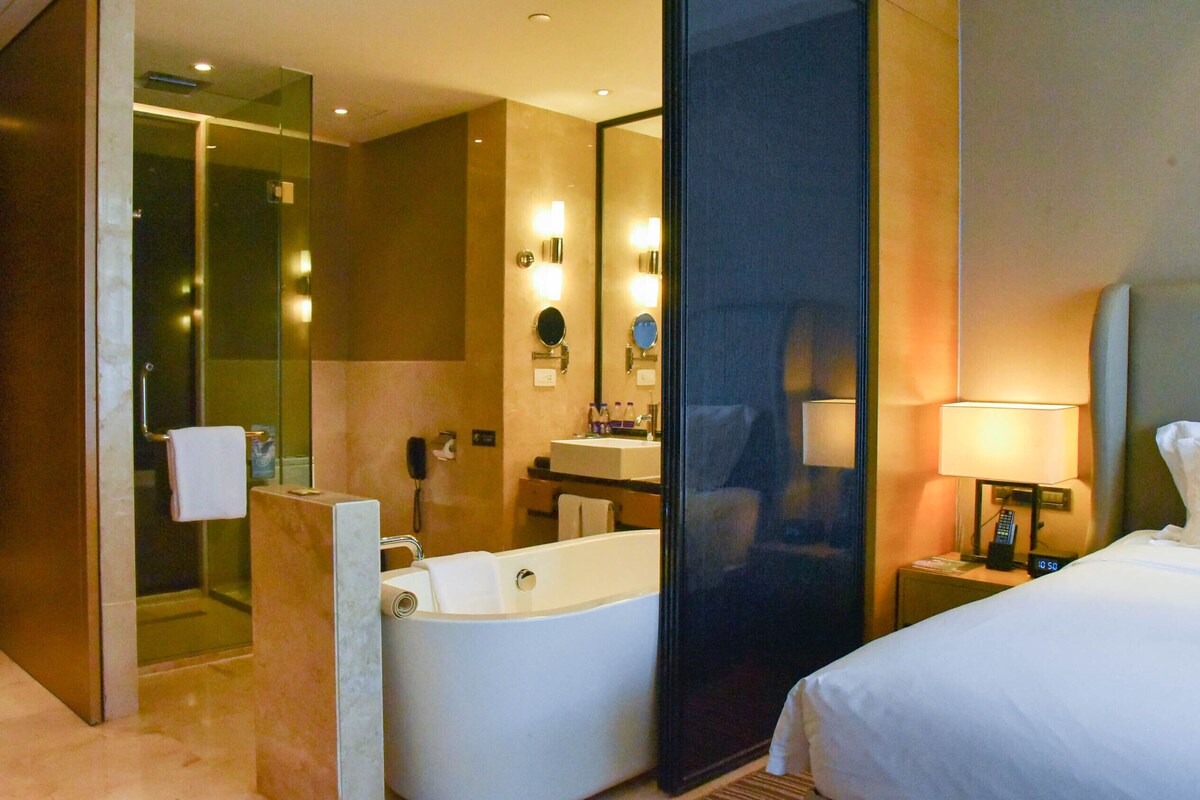Habitación de hotel con una bañera grande, zona de ducha separada, cama y mesita de noche con lámpara.