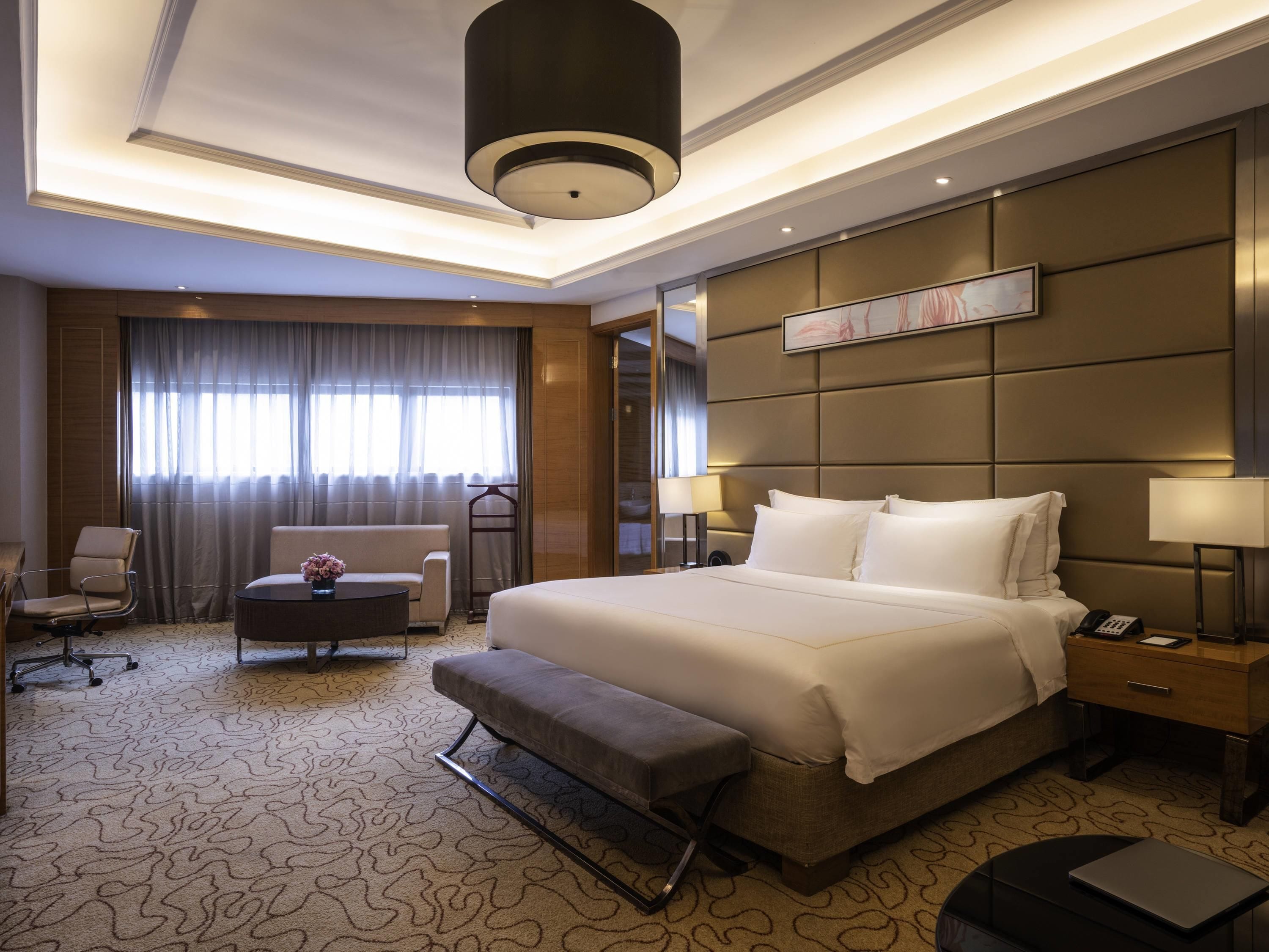 A hotel room with a large bed, a desk, a chair, a bench, a lamp, and a bedside table.