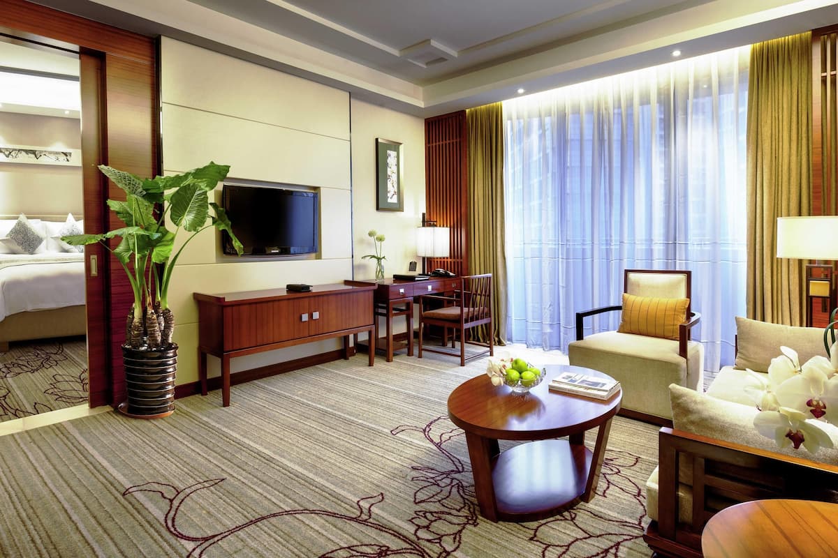 A hotel room with a bed, a TV, a desk, a chair, a round table, and a potted plant.