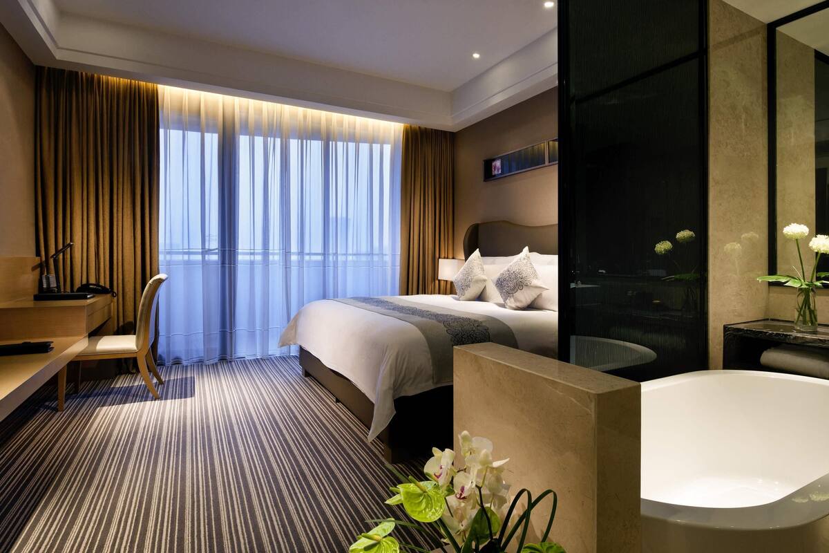 A modern hotel room with a large bed, a desk, a chair, a bathroom with a bathtub, and a large window with curtains.