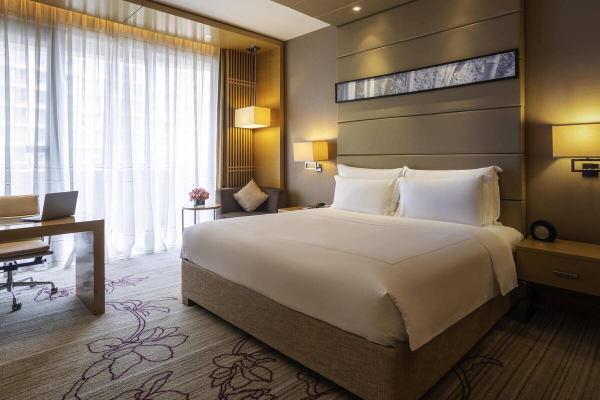 A hotel room with a large bed, a desk with a laptop, a chair, a bedside table with a lamp, and a wall-mounted TV.