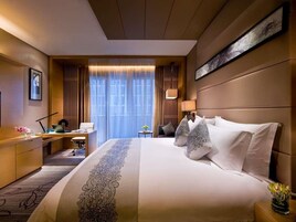 Quarto de hotel com uma cama grande, uma escrivaninha, uma cadeira, um abajur e vista para o exterior pela janela.