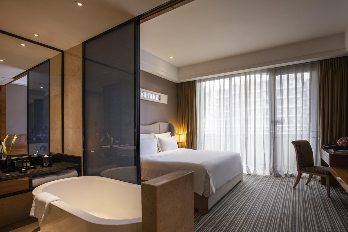 A hotel room with a large bathtub, a bed with white linens, a desk with a chair, and a large window with sheer curtains.
