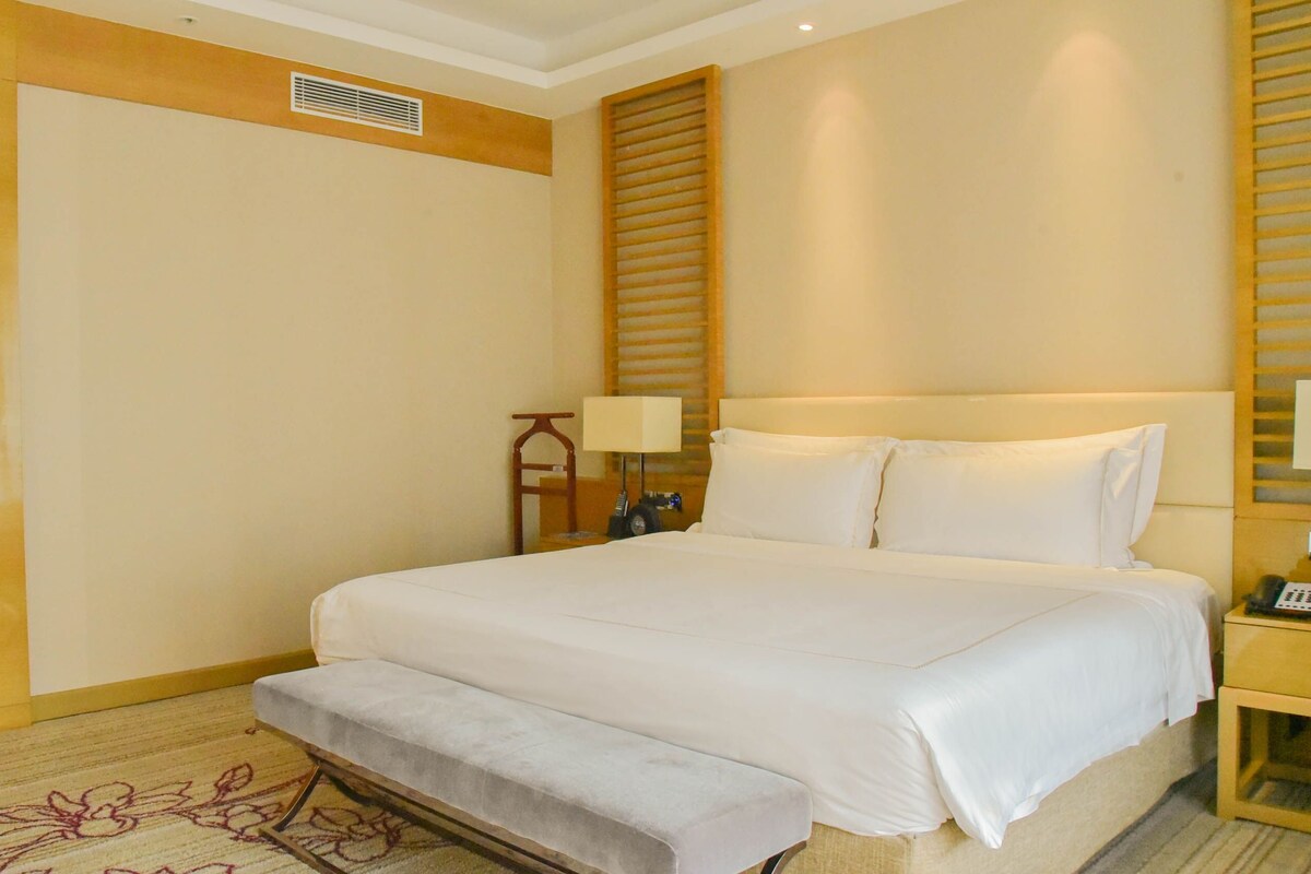 Habitación de hotel con una cama grande, una mesita de noche con lámpara, un banco, un inodoro y un armario.