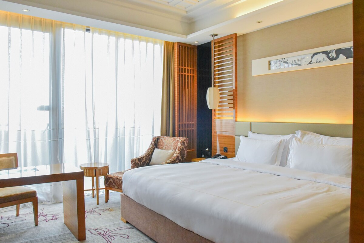 Habitación de hotel con una cama grande, un escritorio, una silla, una mesita de noche y vistas al exterior a través de una ventana con cortinas.