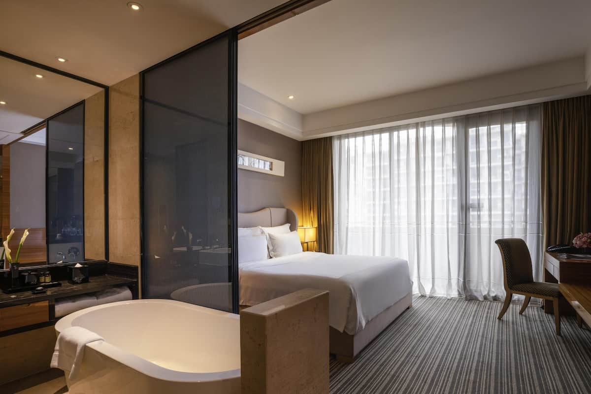 A hotel room with a large bathtub, a bed with white linens, a desk with a chair, and a large window with sheer curtains.