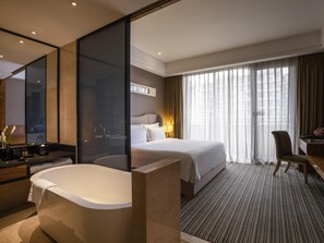 A hotel room with a large bathtub, a bed with white linens, a desk with a chair, and a large window with sheer curtains. - Pullman Foshan Shunde (Foshan)