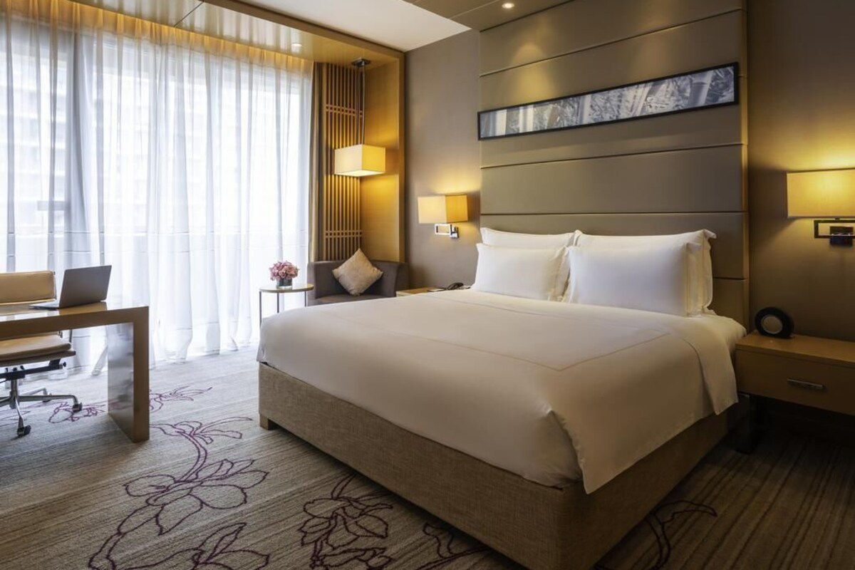 Habitación de hotel con una cama grande, un escritorio con una computadora portátil, una mesita con un florero y un ventanal con cortinas traslúcidas.
