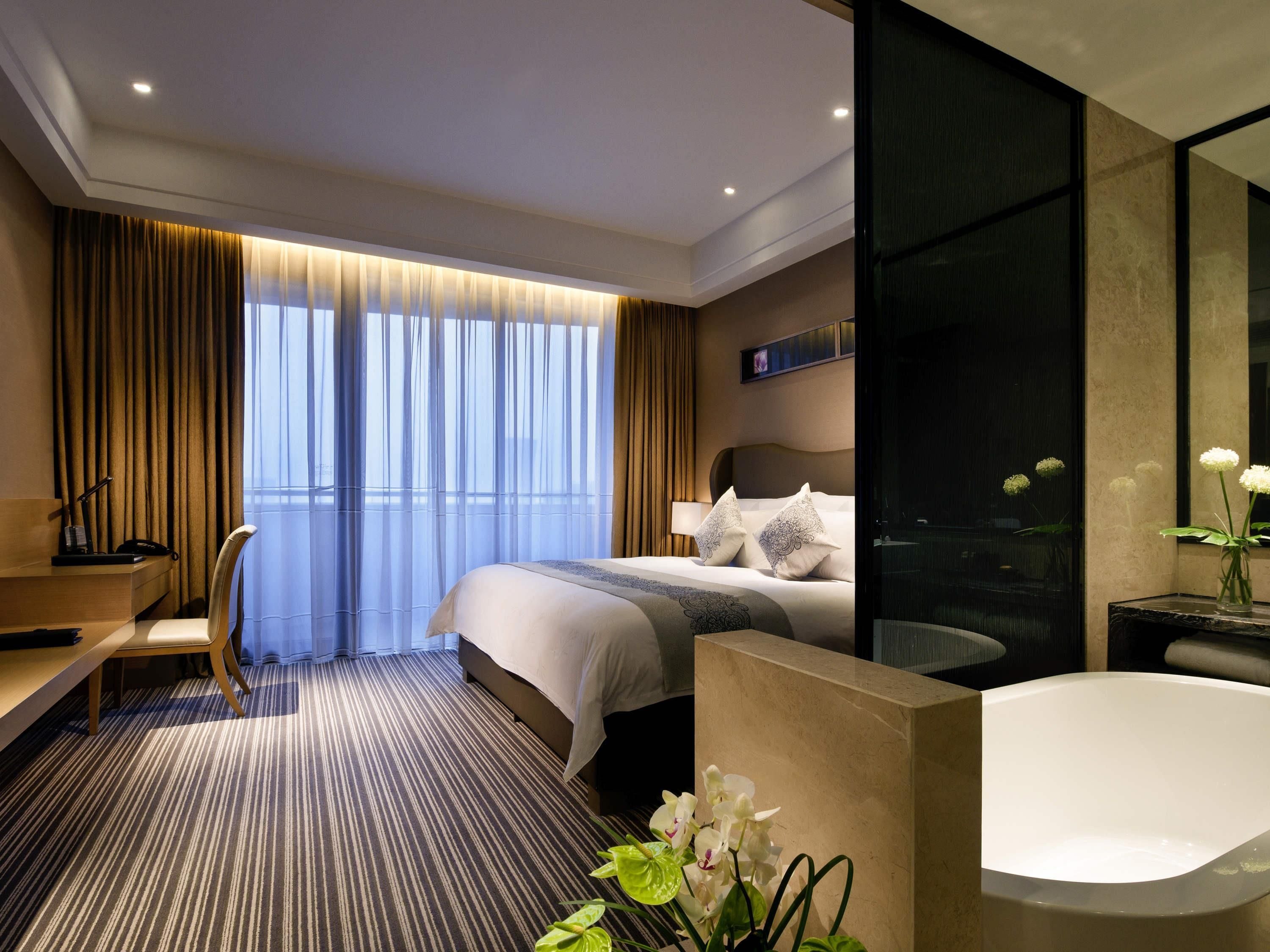 A modern hotel room with a large bed, a desk, a chair, a bathroom with a bathtub, and a large window with curtains.