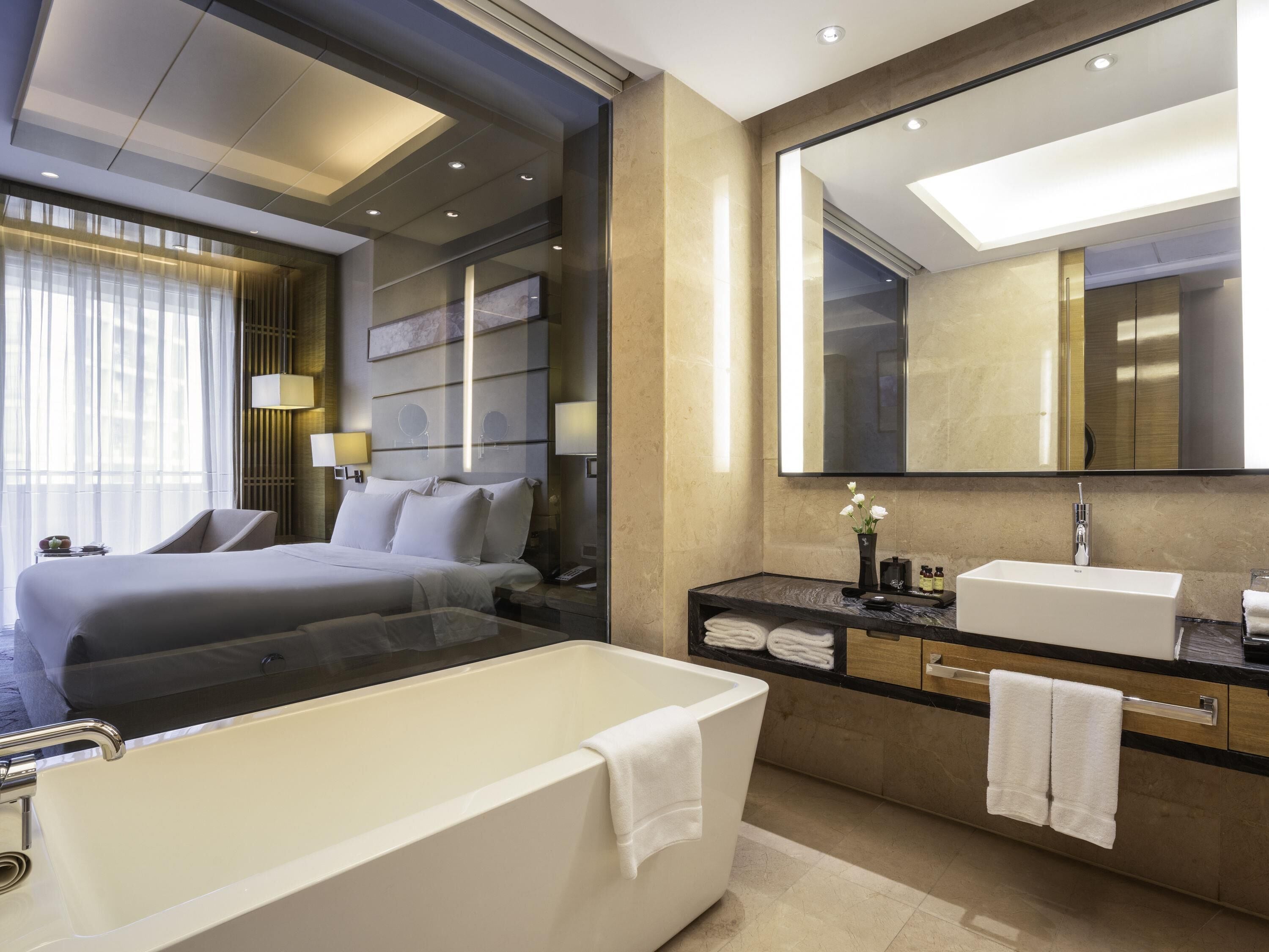 Una moderna camera d'hotel con un letto grande, un'area salotto separata, uno specchio ampio, una vasca e un angolo trucco con un lavandino.