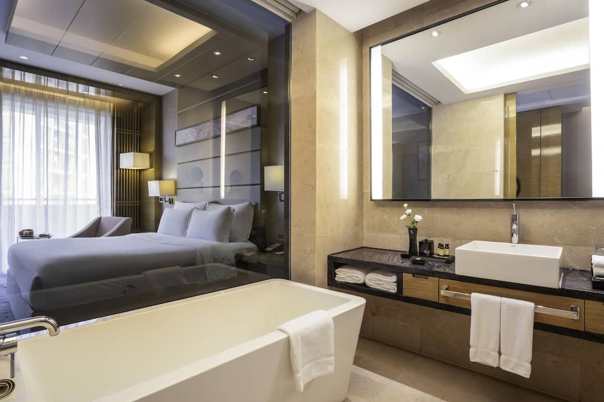 A modern hotel room with a large bed, a separate sitting area, a large mirror, a bathtub, and a vanity with a sink.