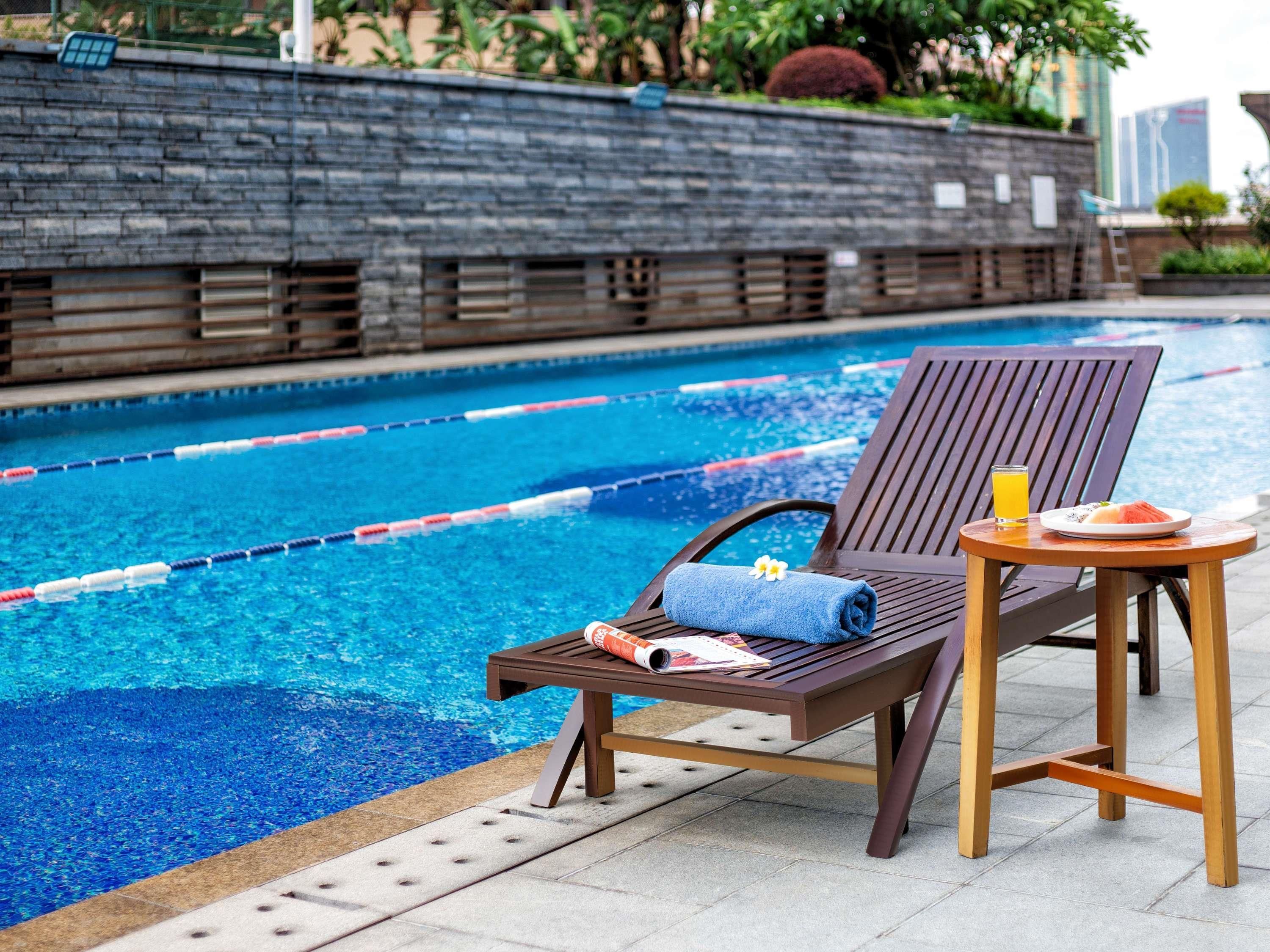 A poolside lounge area with a wooden chair and table, a towel, a glass of orange juice, and a plate of watermelon.