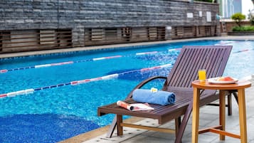 A poolside lounge area with a wooden chair and table, a towel, a glass of orange juice, and a plate of watermelon.