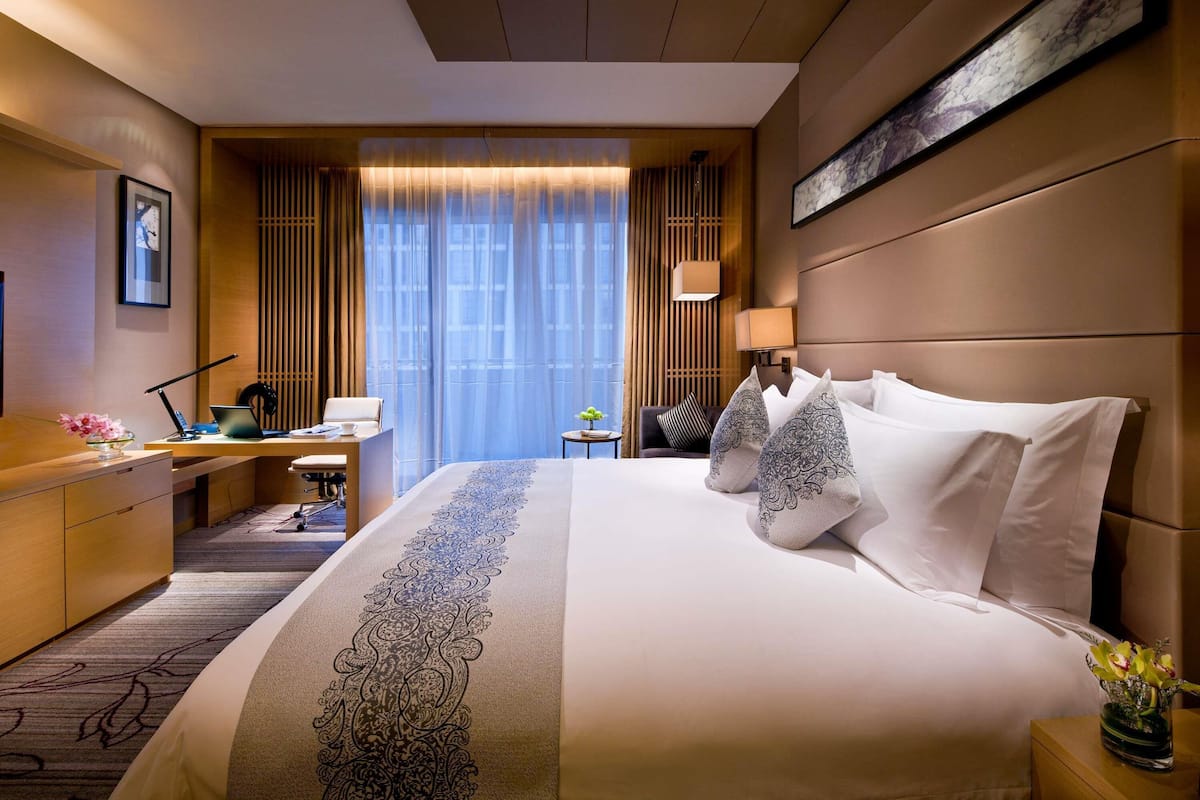 A hotel room with a bed, desk, chair, lamp, and artwork on the wall.