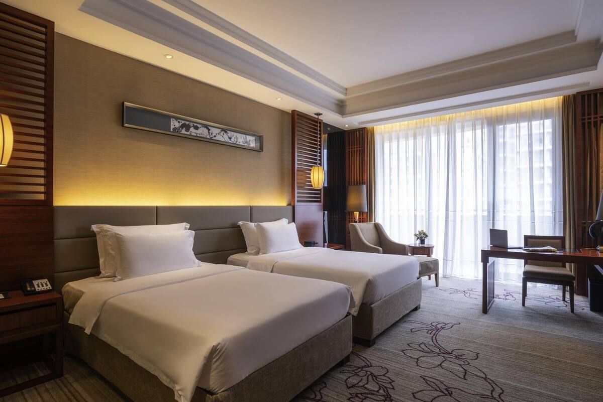 A hotel room with two beds, a desk, a chair, and a large window with curtains.