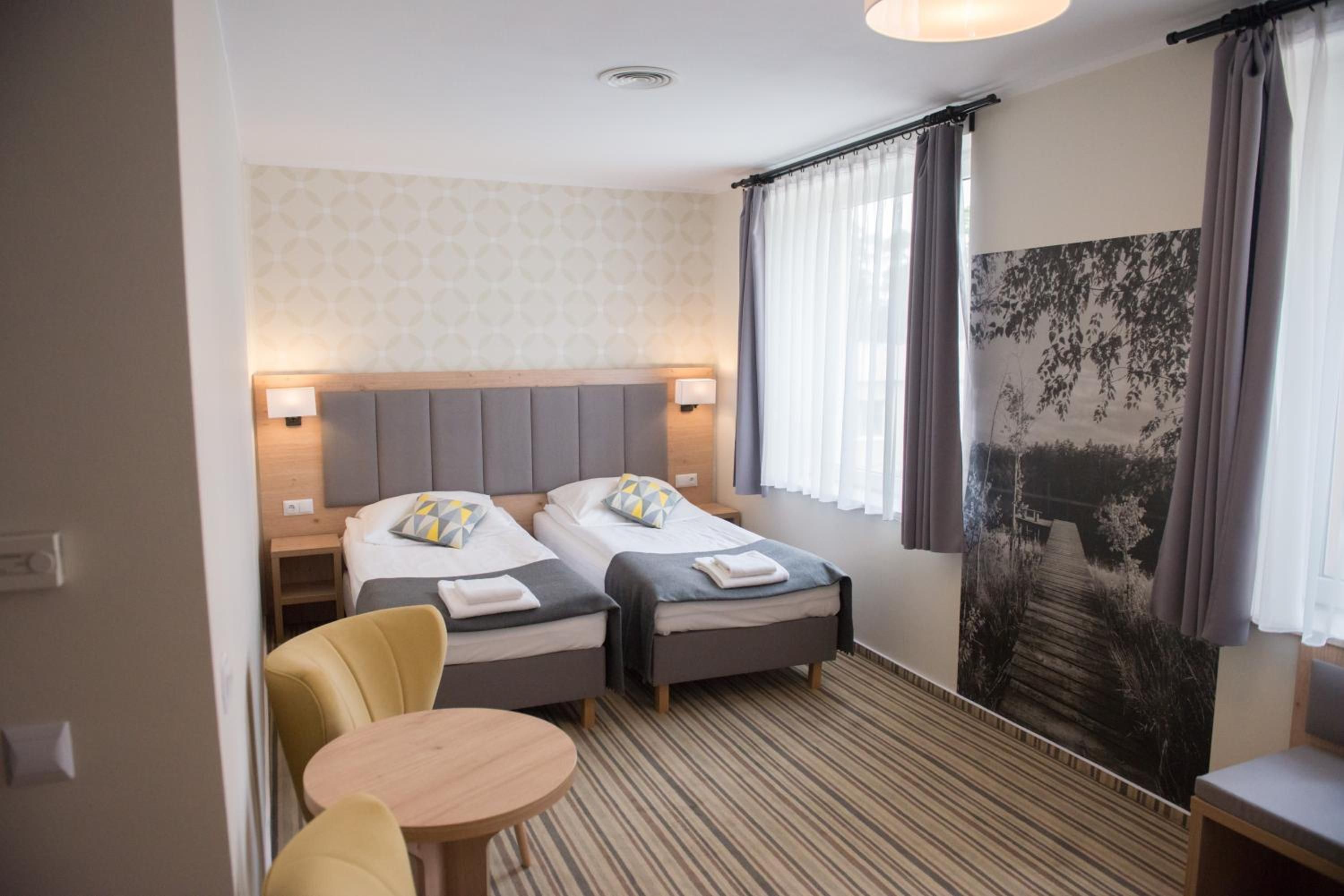 A hotel room with two beds, a small table, a chair, and a large window with curtains.