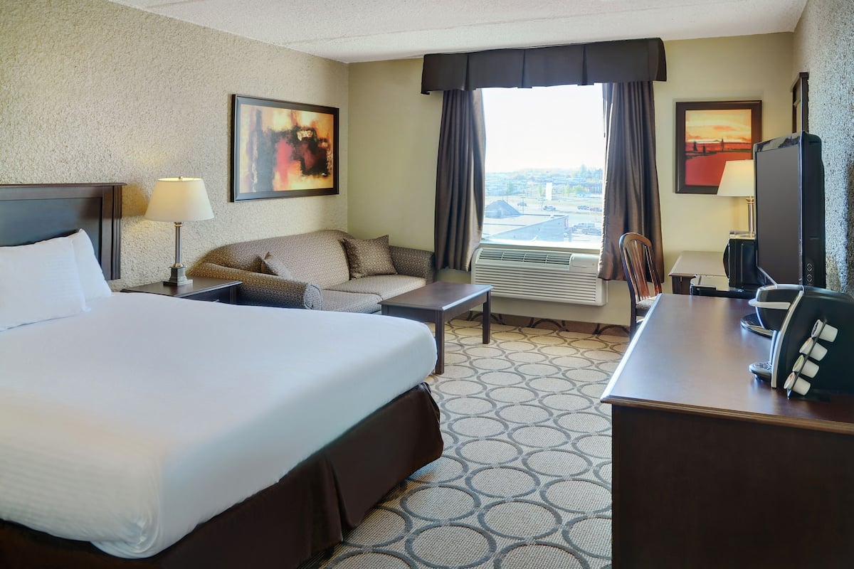 A hotel room with a large bed, a sofa, a desk, and a chair. There are two windows with curtains, an air conditioning unit, and a television.