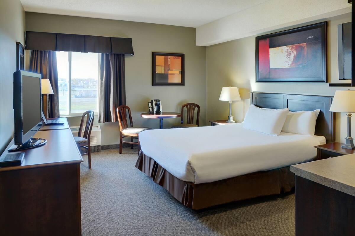 A hotel room with a large bed, a desk with a computer, a chair, a small table with a phone and a coffee maker, and a window with curtains.
