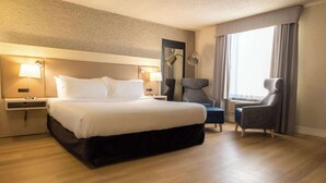 Ein Hotelzimmer mit einem großen Bett, zwei Nachttischlampen, einem Sessel, einem Schreibtisch und einem Fenster mit Vorhängen.