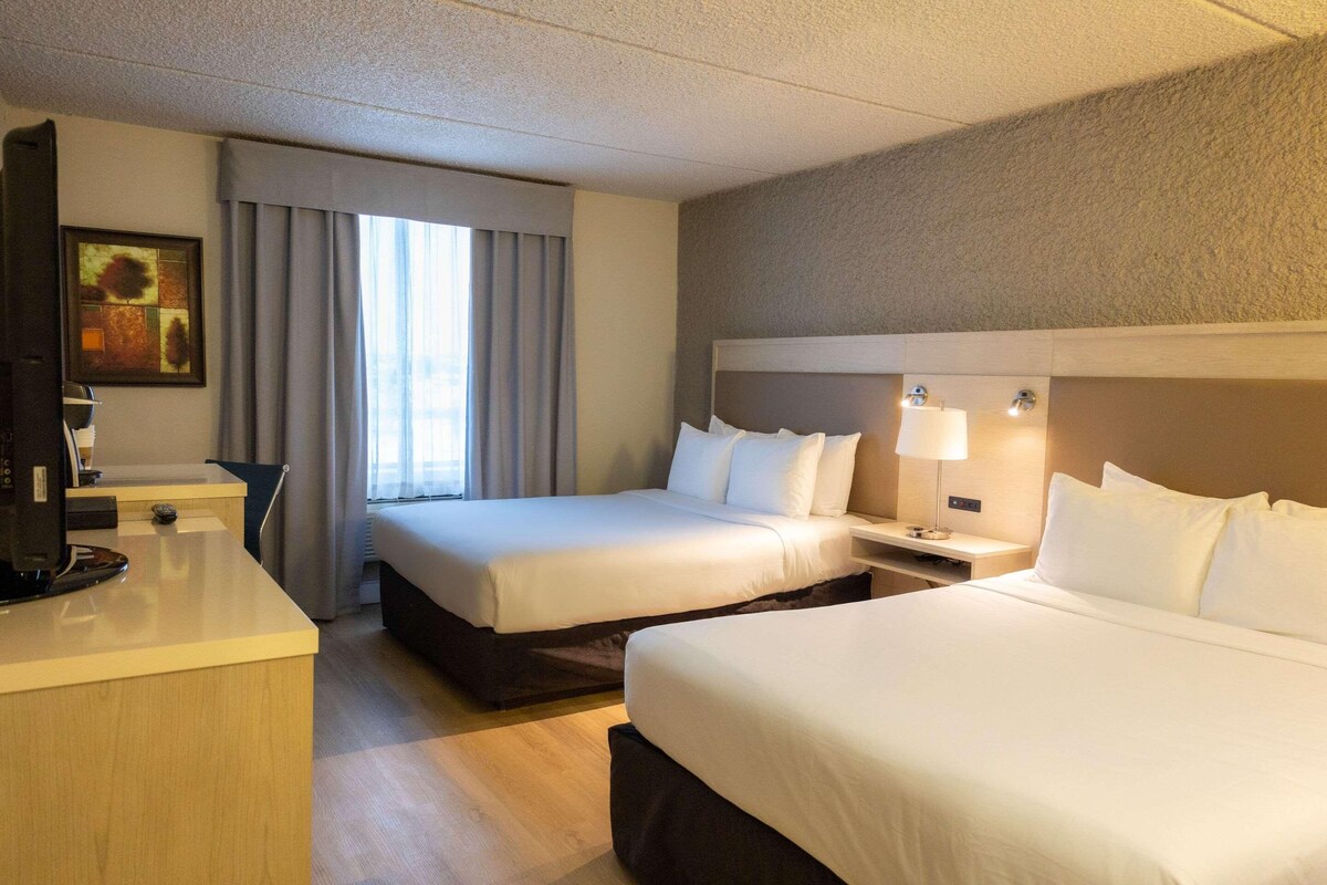 Ein Hotelzimmer mit zwei Betten, einem Schreibtisch, einem Fernseher und einem Fenster mit Vorhängen.