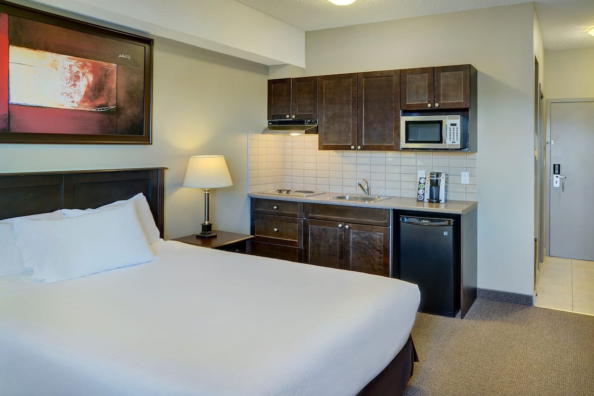 A hotel room with a bed, a nightstand, a lamp, a microwave, a coffee maker, and a small kitchen area.