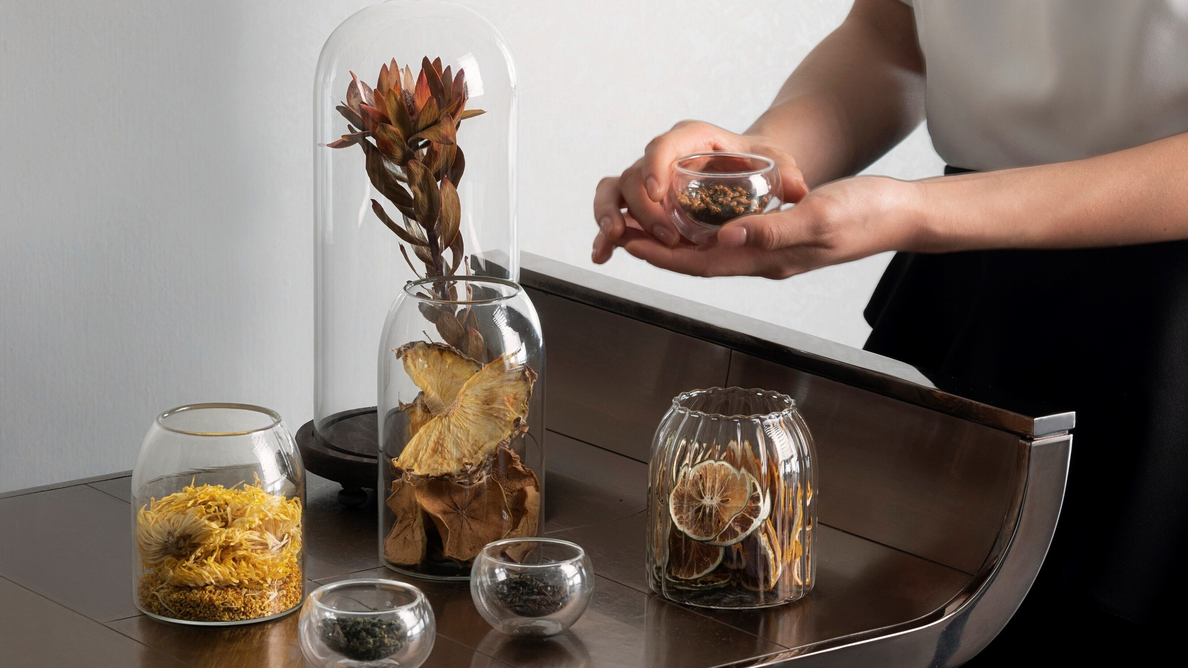 A table with various glass containers holding dried flowers and leaves, a person holding a small bowl, and a clear glass dome over a plant.
