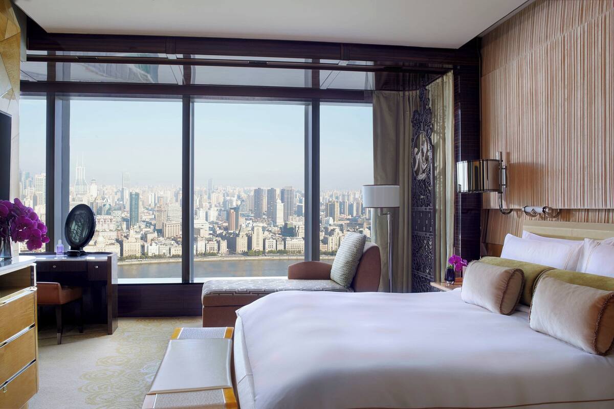A hotel room with a large window offering a city view, a bed with pillows, a desk with a lamp, and a chair.