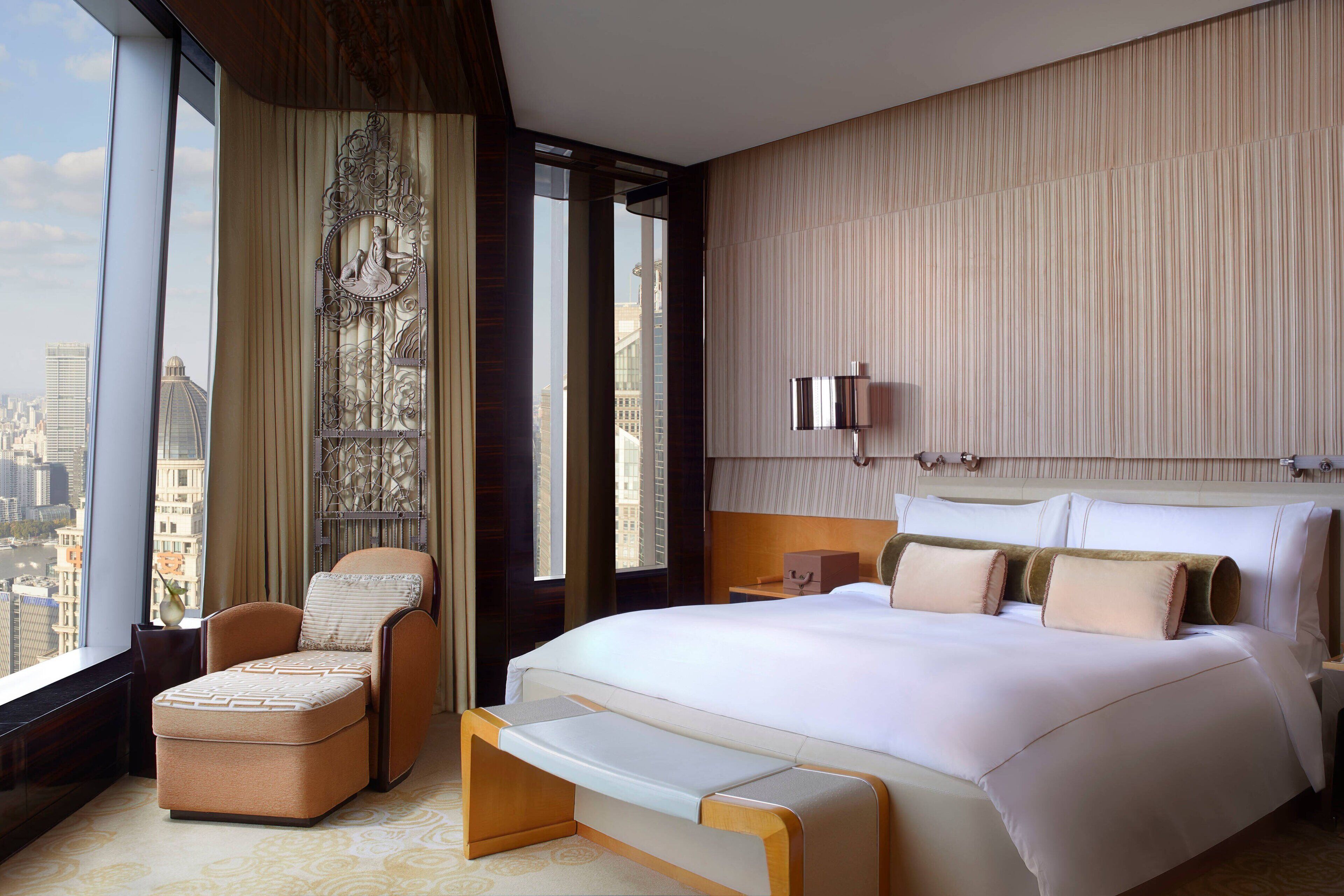 Habitación de hotel moderna con una cama grande, una zona de estar con un banco acolchado, un elemento decorativo en la pared y vistas a la ciudad a través de ventanas amplias.
