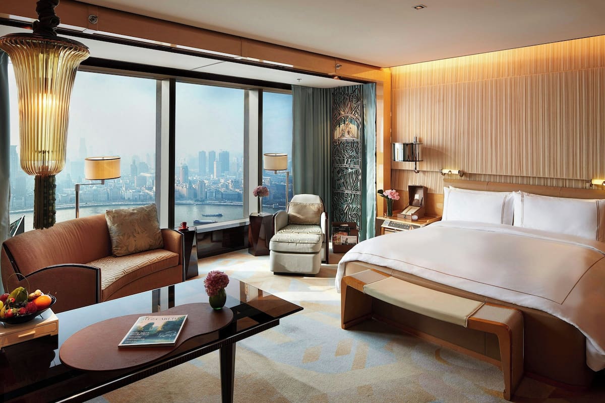 A modern hotel room with a large bed, a sofa, a dining table, and a view of the cityscape.