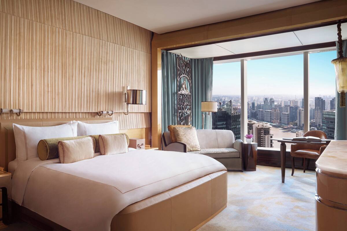 A spacious hotel room with a large bed, a sofa, a dining table, and a view of the cityscape.