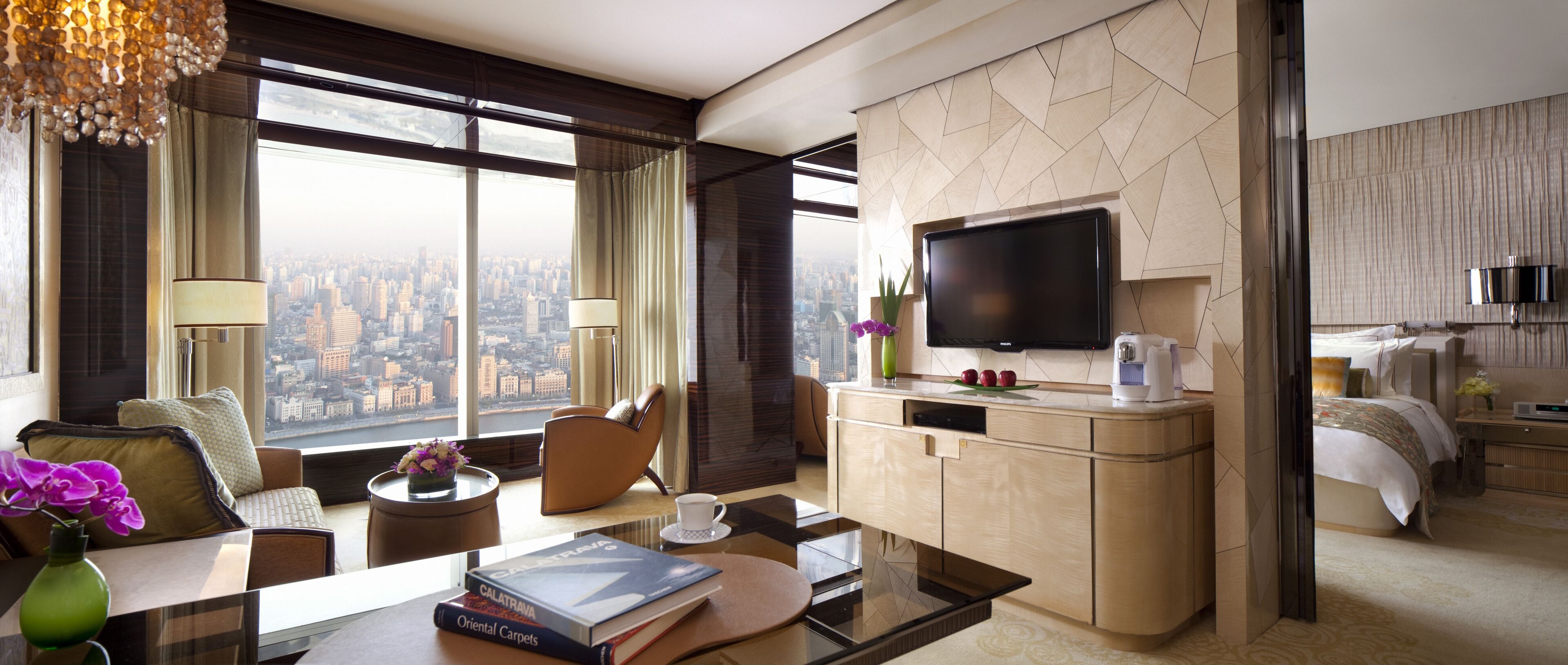 A modern hotel room with a large window offering a city view, a flat-screen TV, a minibar, a desk with a coffee maker, and a comfortable seating area.