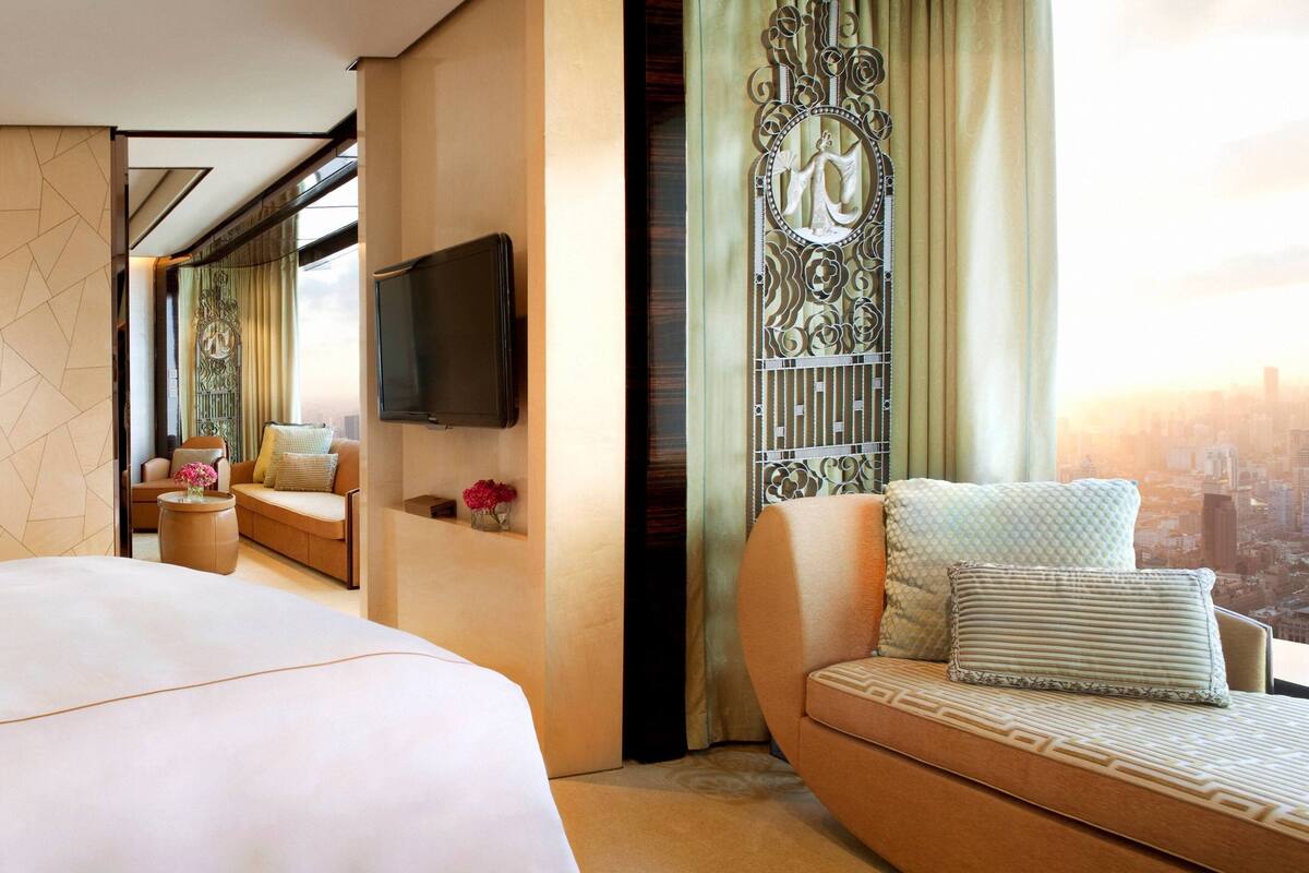 A modern hotel room with a large bed, a sofa, a TV mounted on the wall, and a view of the cityscape.