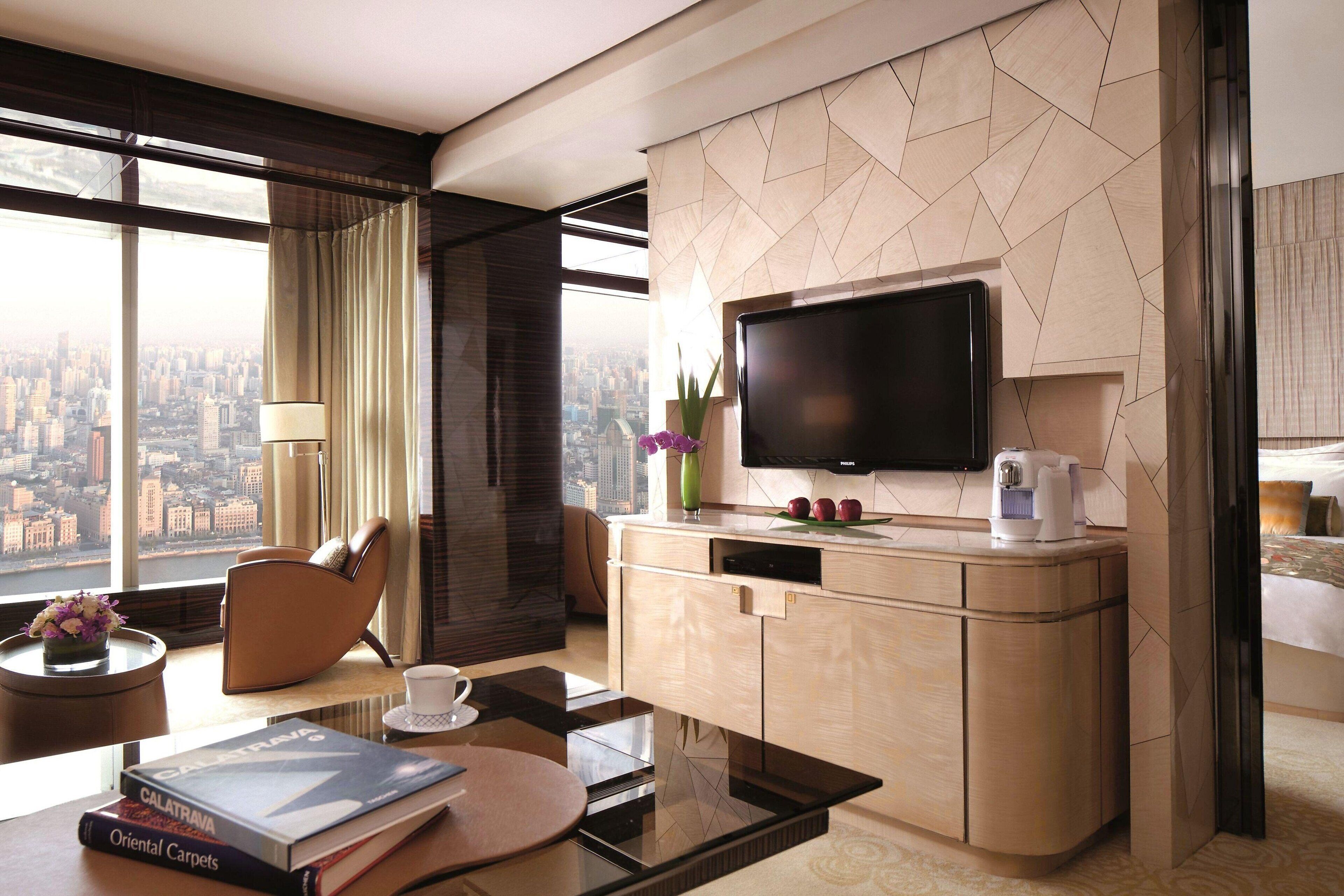 Una habitación de hotel moderna con un ventanal grande, un televisor de pantalla plana, una cafetera, un sofá, una mesa de comedor y una estantería.