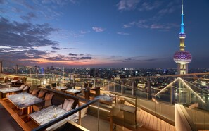 A rooftop bar with outdoor seating, a glass railing, and a city skyline view at dusk.