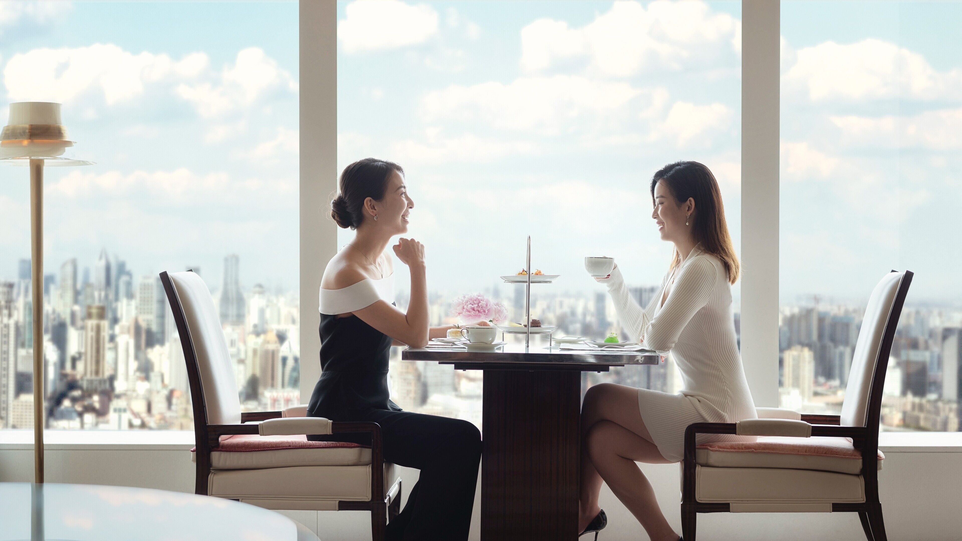 Two people seated at a table with a city view, a dessert tray, and a lamp in a modern room.