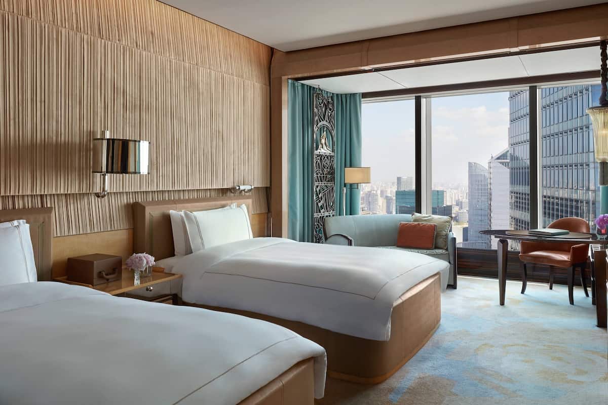 A hotel room with two beds, a desk, a chair, a table, and a large window offering a city view.