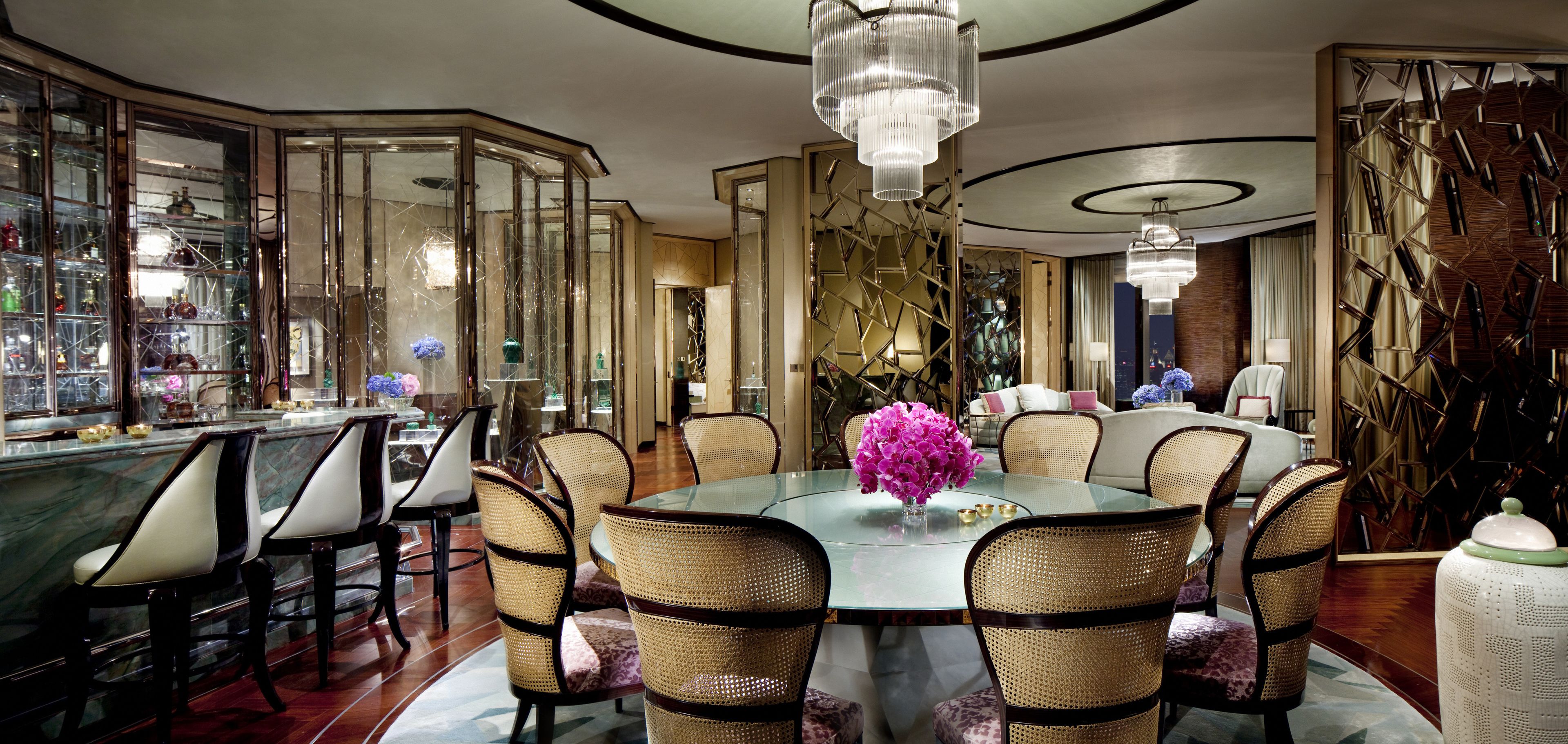 A spacious dining area with a round glass table surrounded by wicker chairs, a bar with glass display cases, and a decorative wall with geometric patterns.