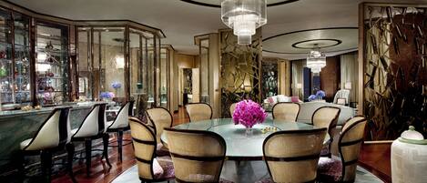 A spacious dining area with a round glass table surrounded by wicker chairs, a bar with glass display cases, and a decorative wall with geometric patterns.