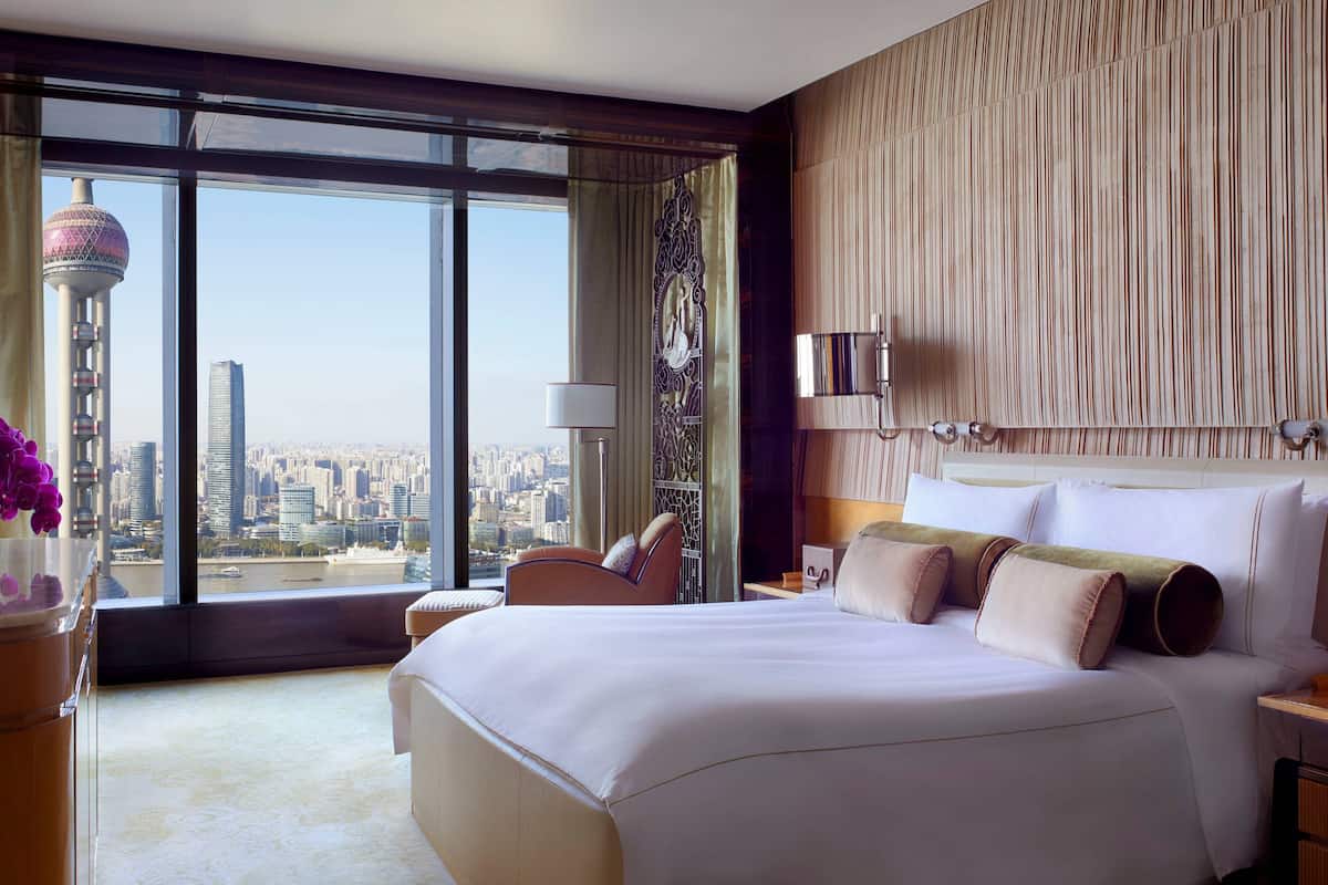 A spacious hotel room with a large bed, a sitting area, and a view of a cityscape through floor-to-ceiling windows.