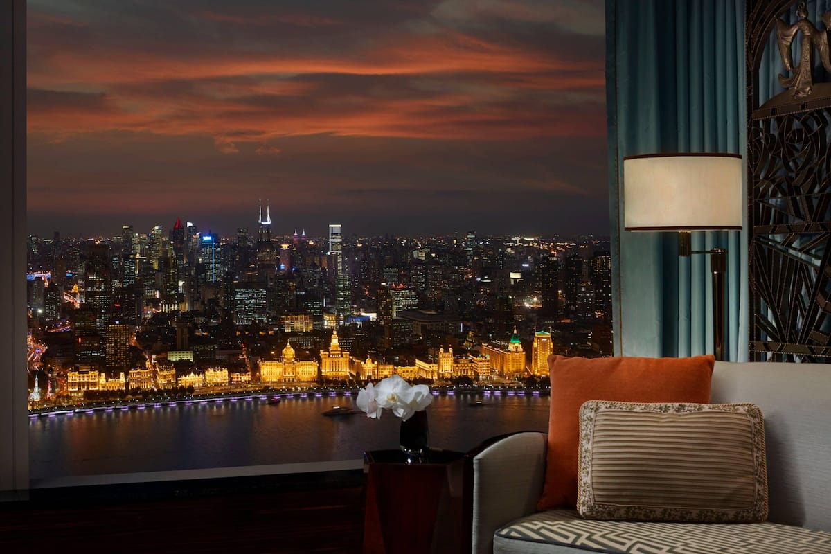 A room with a view of a city skyline at dusk, a sofa with cushions, a table with a vase, and a decorative lamp.