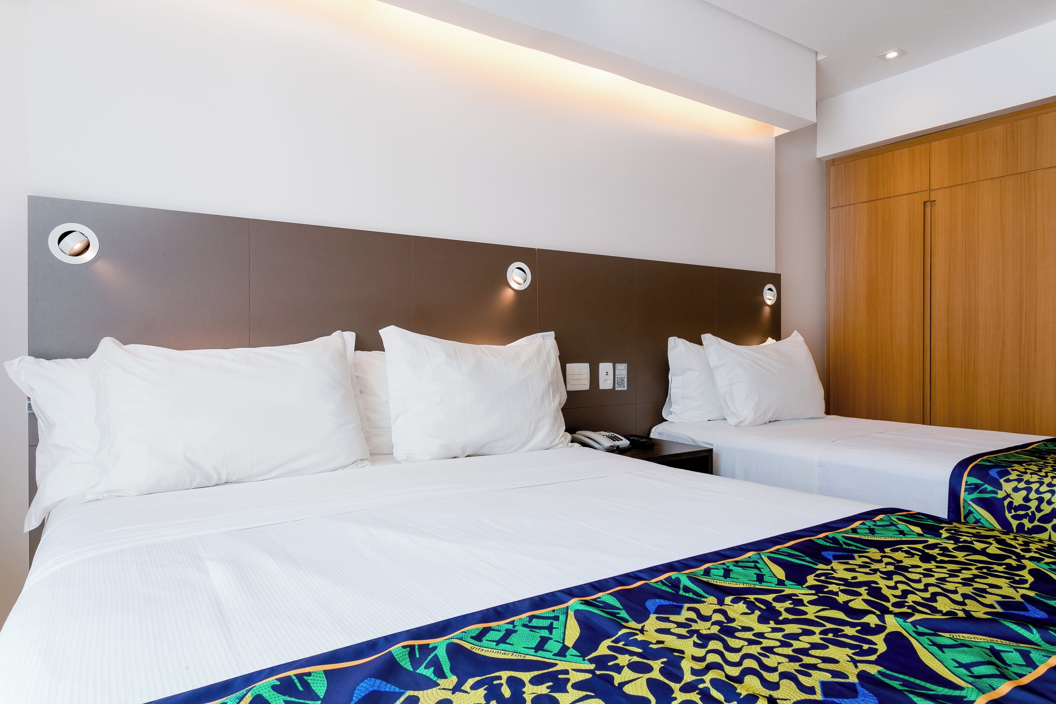 A hotel room with a large bed, two bedside tables, a wooden wardrobe, and a wall-mounted light fixture.