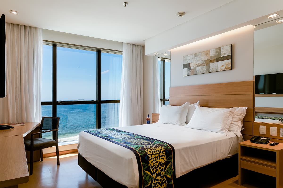 A hotel room with a large bed, a desk, and a chair. There is a window with a view of the ocean.