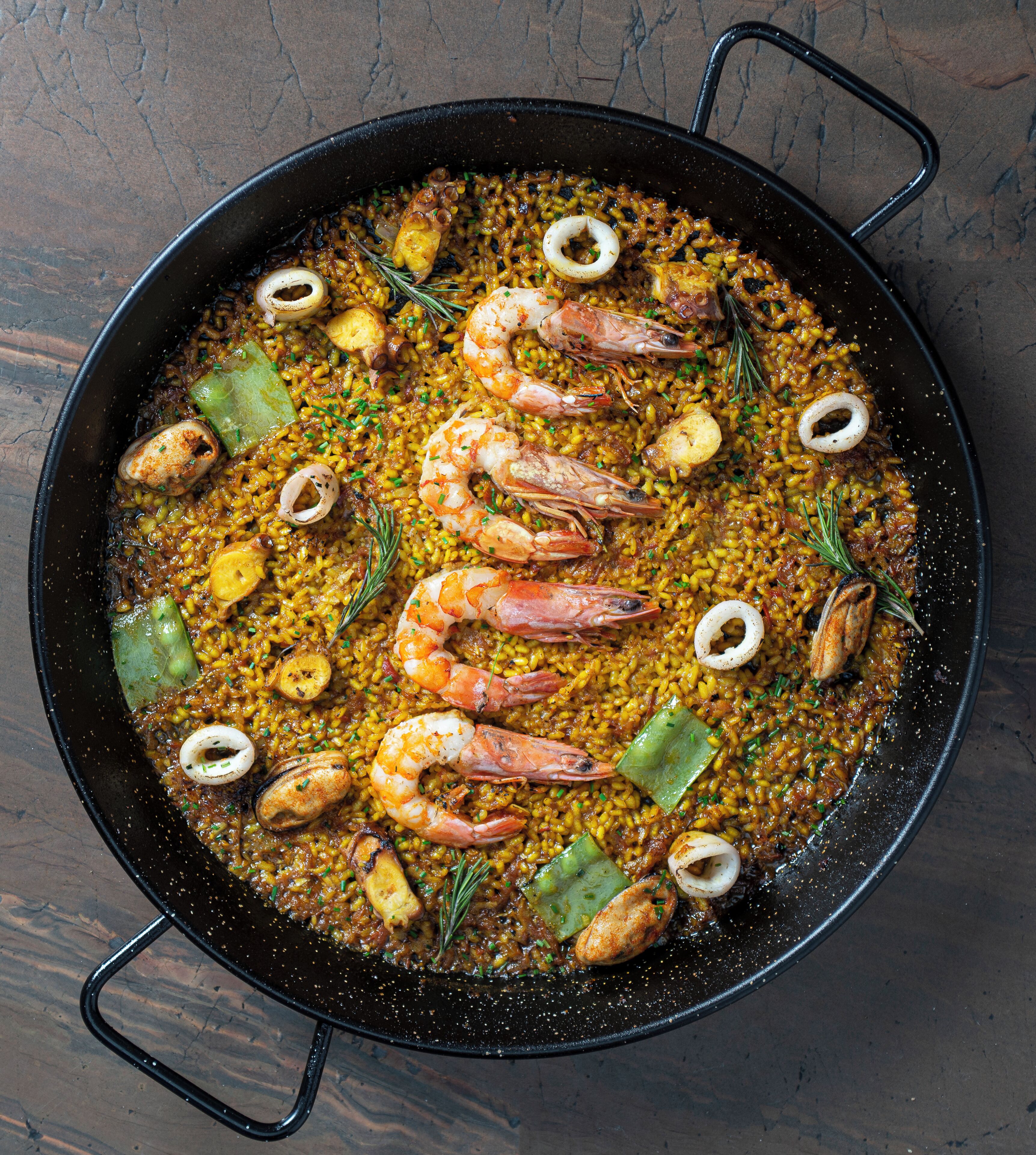A large black pan filled with seafood paella, including shrimp, squid, and rice, garnished with herbs.