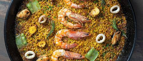 A large black pan filled with seafood paella, including shrimp, squid, and rice, garnished with herbs.
