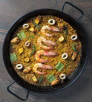 A large black pan filled with seafood paella, including shrimp, squid, and rice, garnished with herbs.