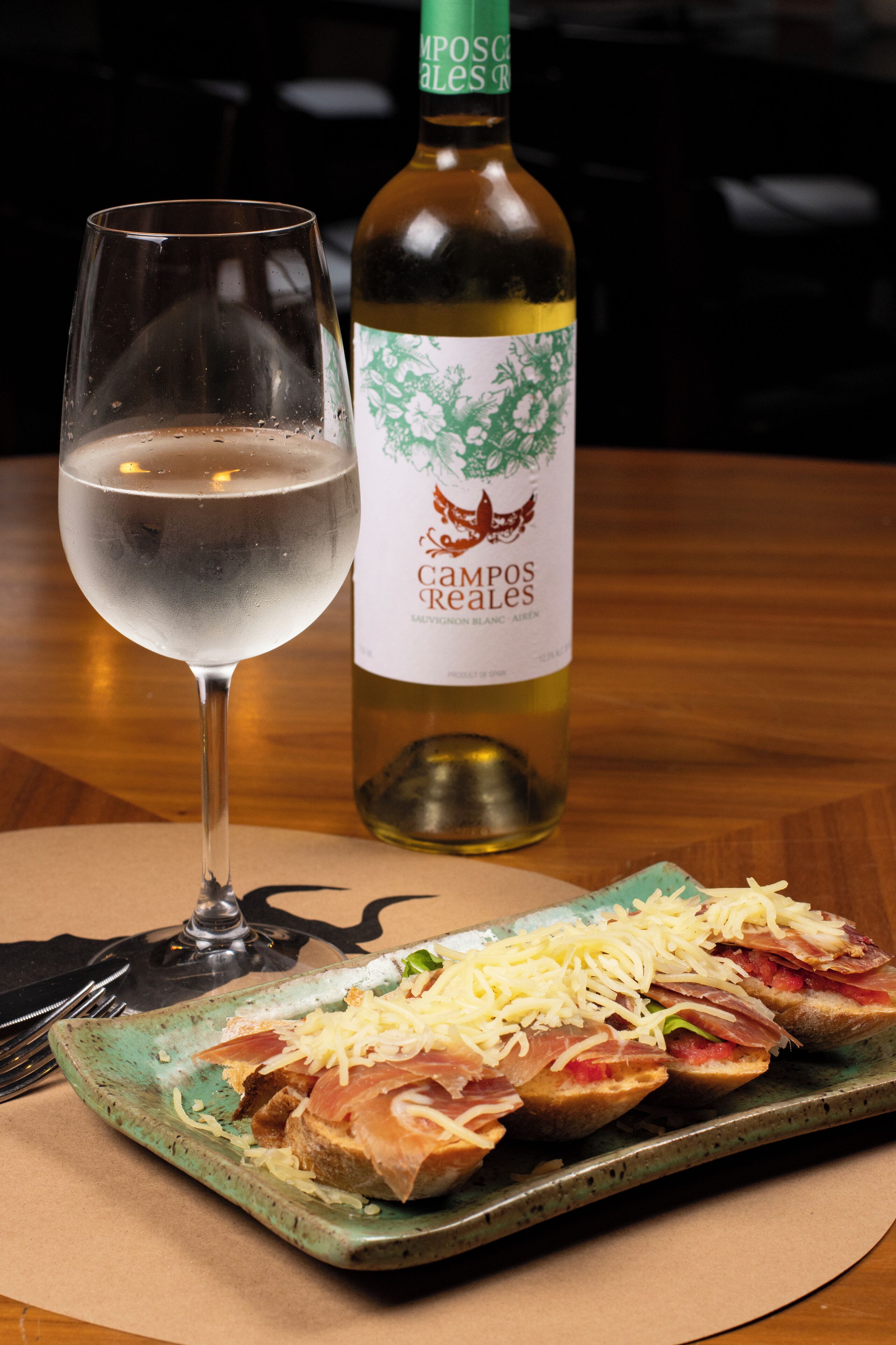 A glass of white wine, a bottle of Campos Realés, and a plate of bruschetta with cheese and prosciutto.