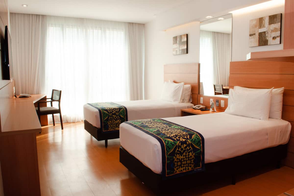 A hotel room with two beds, a desk, a chair, a TV, and a large window with curtains.