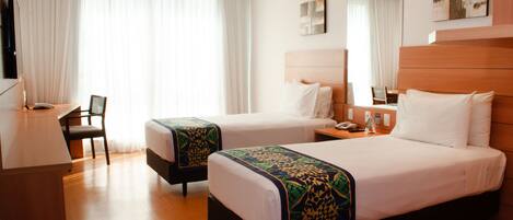 A hotel room with two beds, a desk, a chair, a TV, and a large window with curtains.